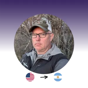 A man with a baseball hat and glasses looks at the camera. US flag points to the Argentine flag.