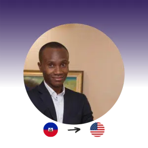 A man in a black suit smiles at the camera. The Haitian flag points to the U.S. flag.